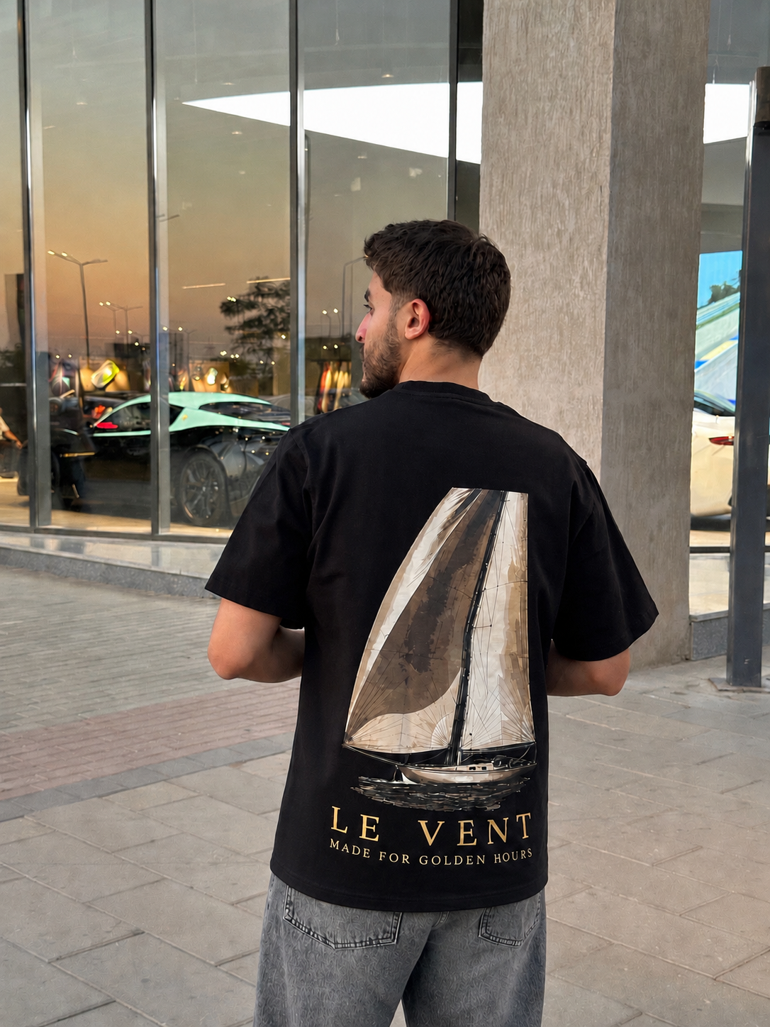 Man in the black AUREO Le Vent oversized tee at golden hour, sailboat back print facing camera